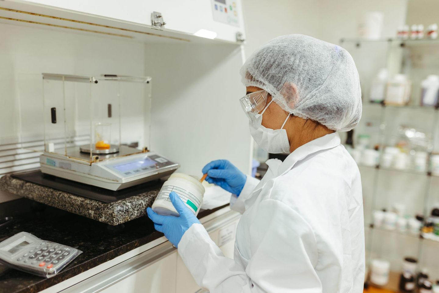 A laboratory technician in a hairnet, mask and gloves weighing a sample on an analytical balance